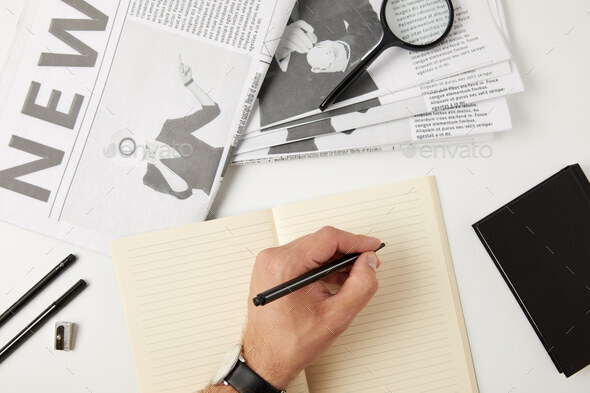 partial top view of person writing in blank notebook, newspapers, magnifying glass, notebook and