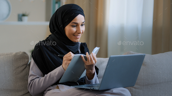Cheerful young muslim arabian lady young woman sit on cozy sofa female freelancer writer take notes