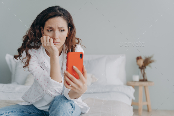 Stressed caucasian woman is doom scrolling. Girl reading morning news on smartphone in her bedroom.