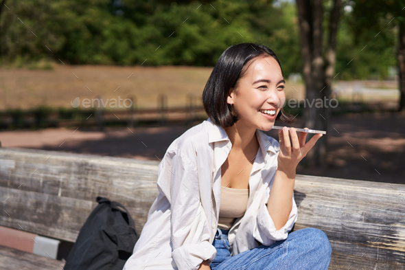 Portrait of happy asian girl records voice message, talking via speakerphone on smartphone, sitting