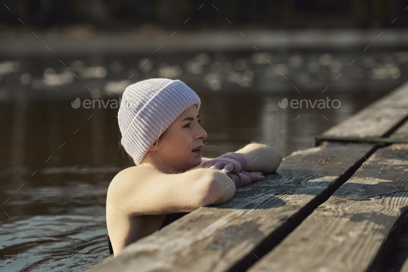 Caucasian adult woman swimming in frozen lake and leaning on wooden pier - Stock Photo - Images