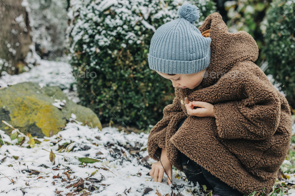 Little kid in hat and coat in winter snowy garden - Stock Photo - Images