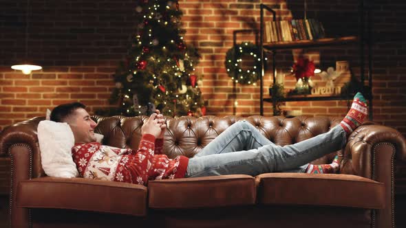 Happy Student Jumping on Couch in Decorated Apartment in Evening After Work Using Mobile Phone