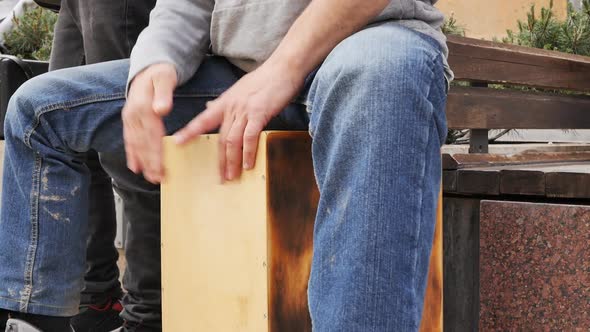 A Street Musician Plays with His Hands on a Cajon While Sitting on It alt