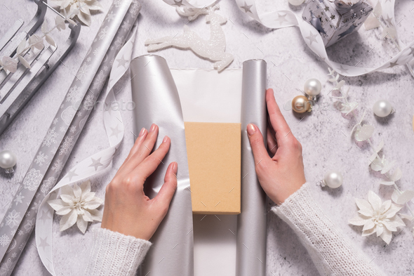 Women's hands are wrapping a Christmas present in silver wrapping paper.  Stock Photo by AllaRudenko