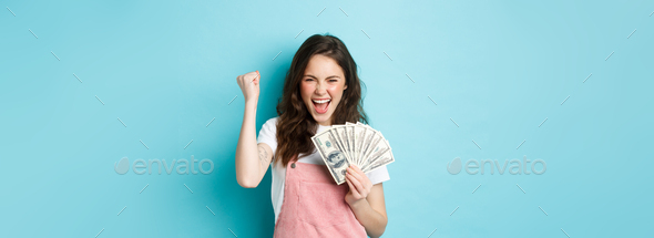 Lucky young woman looks excited, shouting from satisfaction and triumph, winning money, holding