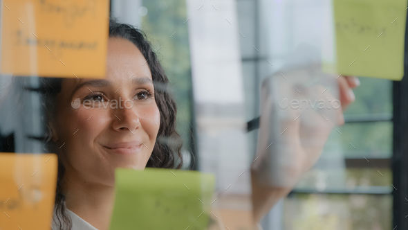 Young positive hispanic woman office worker writes project tasks on sticky notes organizes work