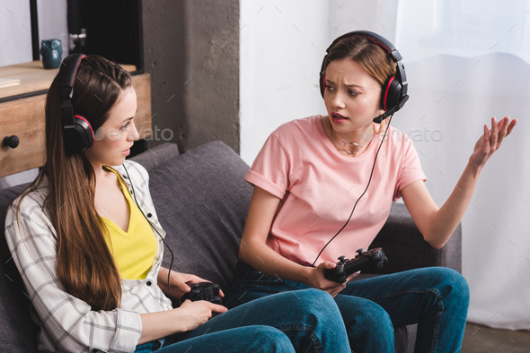angry young woman in headphones with joystick in hand quarreling with female friend at home