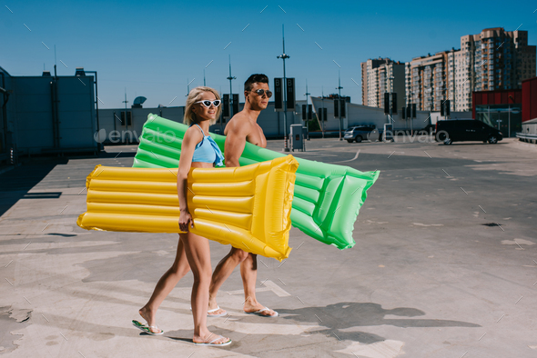 young couple in beach clothes with inflatable beds walking on parking