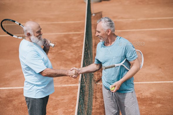 side view of smiling elderly friends with tennis racquets shaking hands after game on court