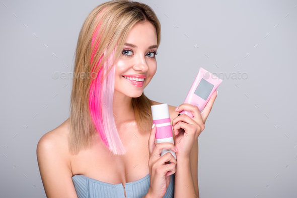 smiling young woman with colored bob cut holding hair care supplies and looking at camera isolated