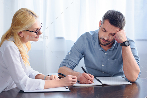 man writing down in empty textbook while sitting at table near female counselor in office - Stock Photo - Images