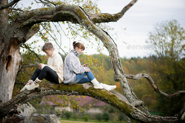 mother and son teen sit together on a tree branch and use cell phones and headsets at city park