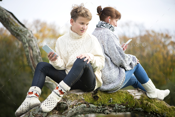 Happy mother and son teen sit with their backs together on a tree branch and use cell phones