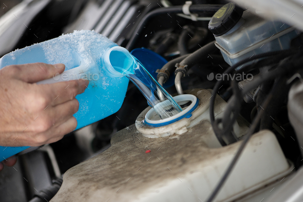 Close up driver pouring blue non freezing windshield glass washer fluid into the tank of the car.