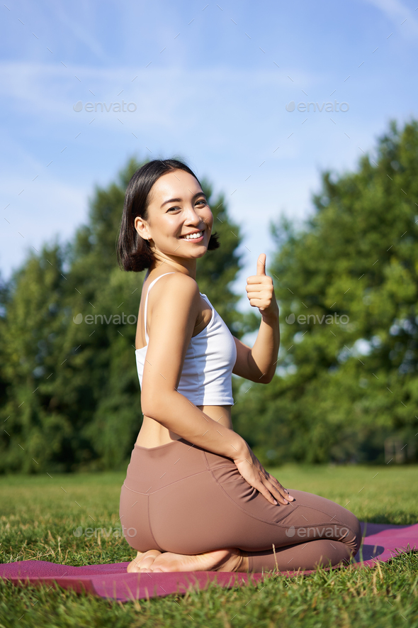 Portrait of asian woman say yes to yoga training in park, makes thumb up, sitting and meditating