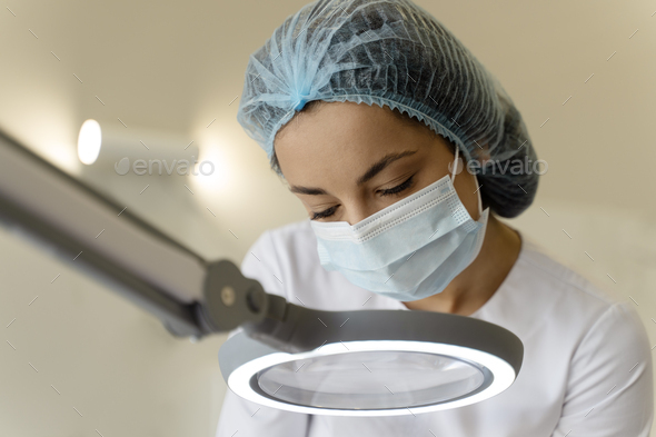 Cropped female cosmetologist looking at client\'s face through magnifying lamp