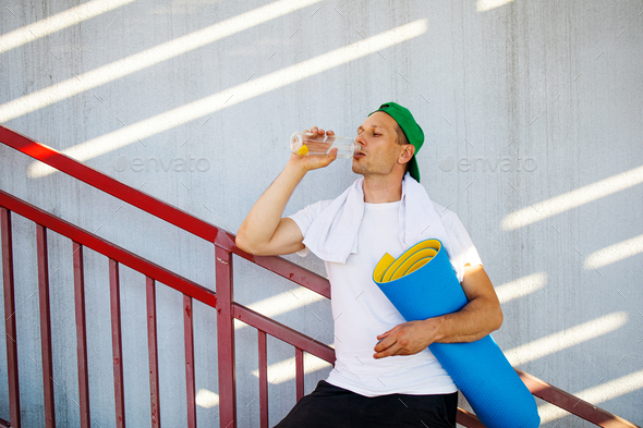 Fitness sport man on stairs drinking water relaxing post-workout in day light