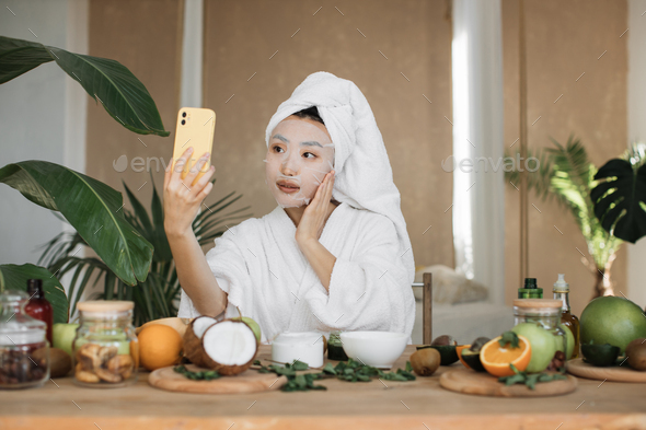 Close up portrait of happy asian woman blogger holding phone applying white cotton mask sheet
