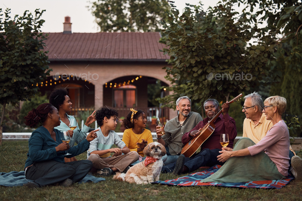 Senior black man playing acoustic guitar while relaxing with his multiracial family in the backyard.