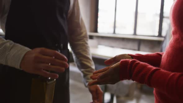 Midsection of man working at cafe bar, using sanitising spray on hands of female colleague alt