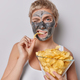 Horizontal shot of European woman with short hair eats delicious crisps enjoys harmful food winks - PhotoDune Item for Sale