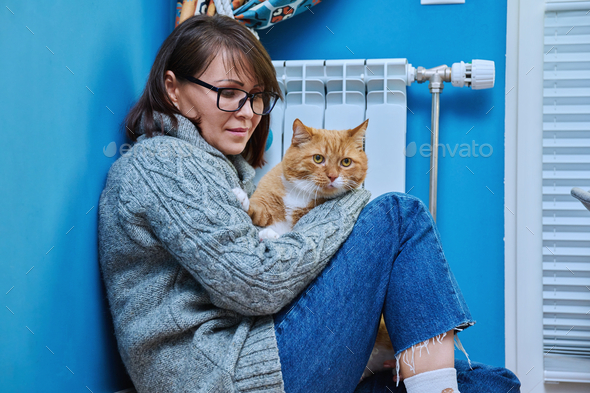 Woman in knitted woolen sweater with pet cat warming near heating radiator - Stock Photo - Images