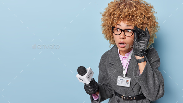 Photo of amazed curly haired woman keeps hand on rim of spectacles prepares reportage piece for