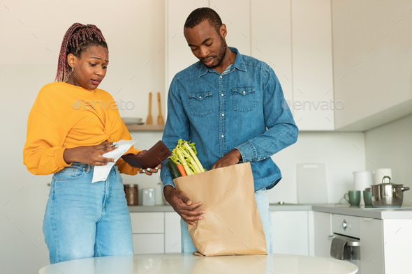 Black couple unpacking products from market in kitchen, woman looking at bill and wallet with shock