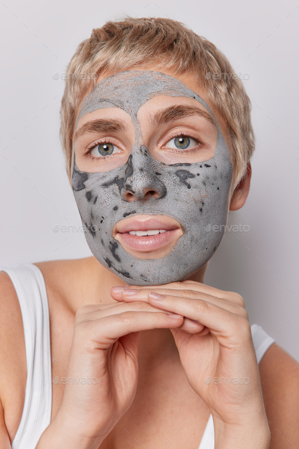 Headshot of beautiful woman with big green eyes applies facial clay mask for reducing fine lines and