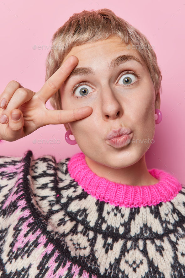 Vertical shot of surprised short haired woman keeps lips rounded makes peace gesture over eye looks