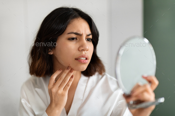 Upset young woman looking at mirror and touching her face - Stock Photo - Images