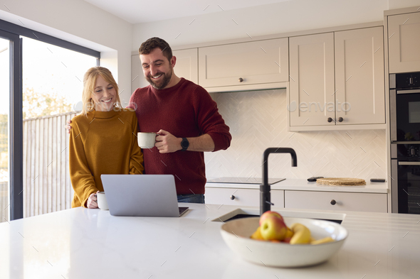 Couple At Home Looking At Laptop On Counter In Kitchen Together
