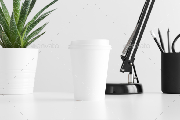 Coffee paper cup mockup with a cactus in a pot, lamp and workspace accessories on a white table.