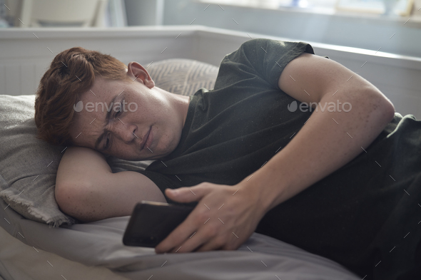 Caucasian teenage boy browsing phone while lying on bed - Stock Photo - Images