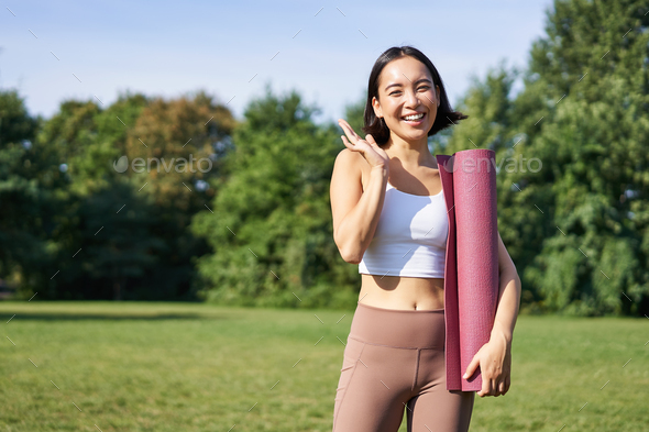 Happy asian fitness girl says hi, waves hand, stands with rubber yoga mat for workout in park