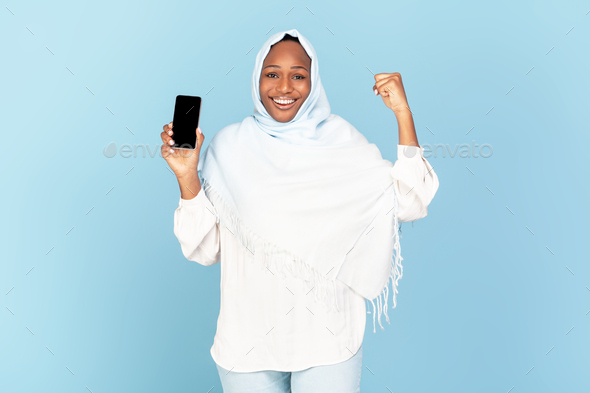 Big luck. Excited black muslim woman holding blank smartphone and shaking fist, exclaiming with