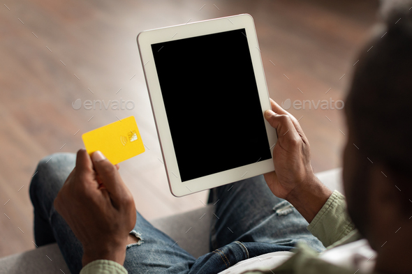 Mature african american man on sofa watch video on tablet with empty screen, show credit card in