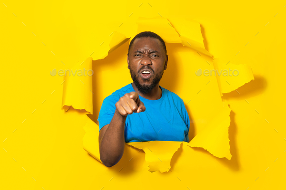 Aggressive black man pointing finger looking at camera, blaming you, posing in hole in torn yellow