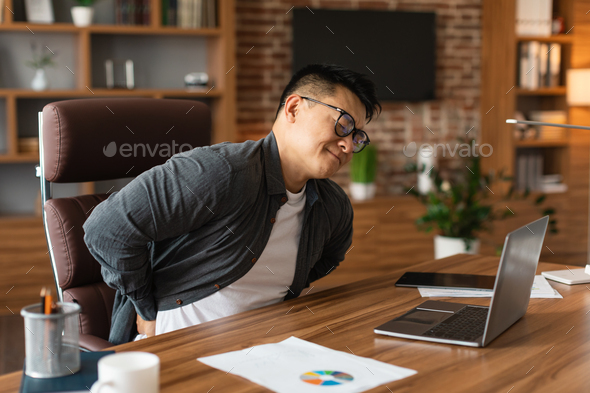 Unhappy middle aged asian man manager in glasses suffering from muscles back pain at table with