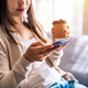 Young woman with cup of coffee using mobile phone and relaxing on sofa in living room at cozy home - PhotoDune Item for Sale