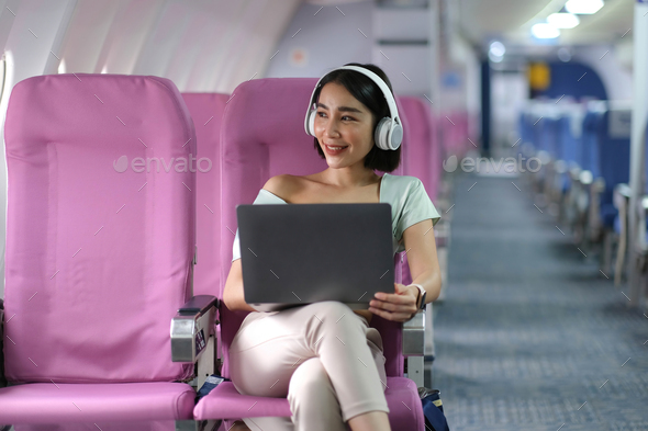 Asian young woman wearing headphone using laptop sitting near windows at first class on airplane - Stock Photo - Images