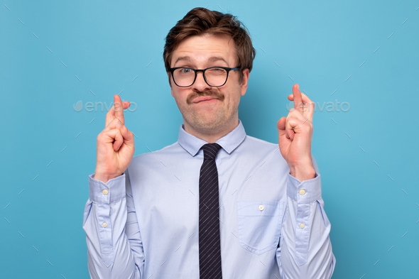 Young caucasian businessman in funny glasses making a wish isolated on blue background.