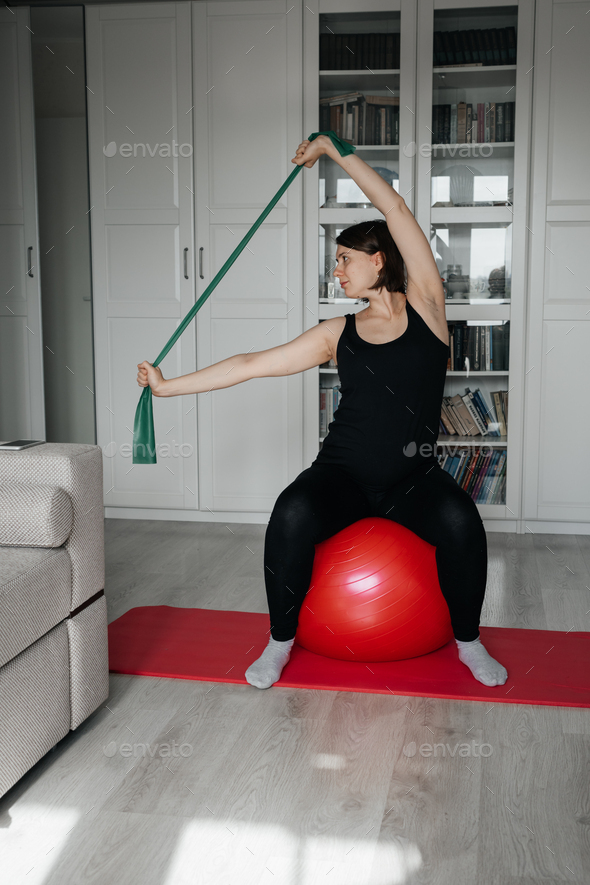 Adult pregnant woman doing exercises with big red fit ball and rubber resistance band at home.