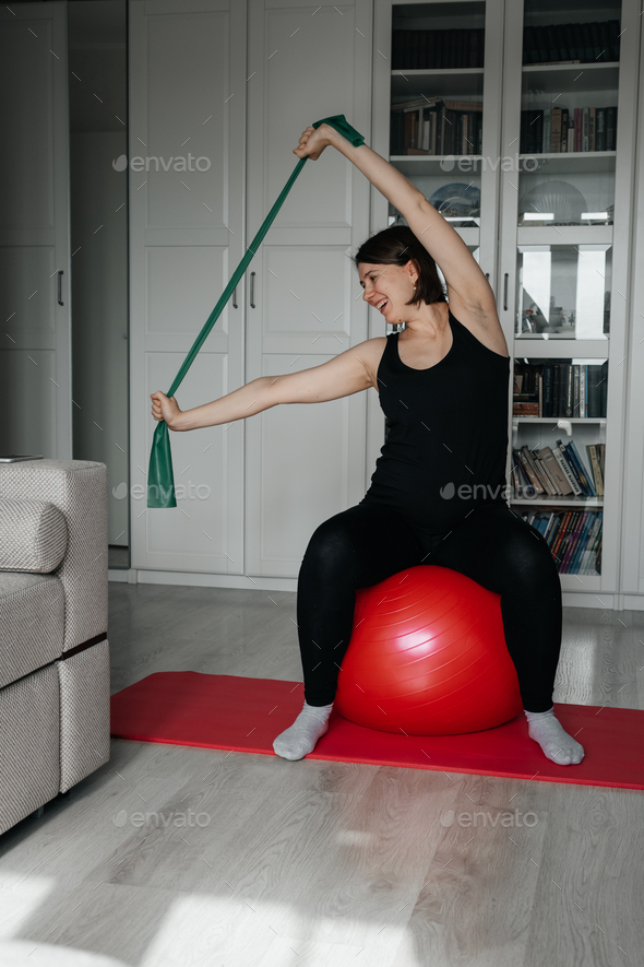 Adult pregnant woman doing exercises with big red fit ball and rubber resistance band at home.