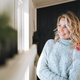 Smiling woman forty year with blonde curly hair in grey knitted sweater near window at house - PhotoDune Item for Sale
