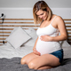 Beautiful pregnant woman sitting on bed resting and touching her belly - PhotoDune Item for Sale