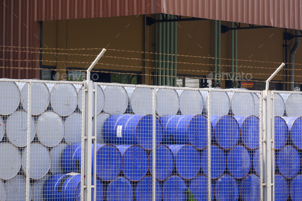 Many blue and white 200 liter chemical or oil drum stacking behind mesh fence in factory area