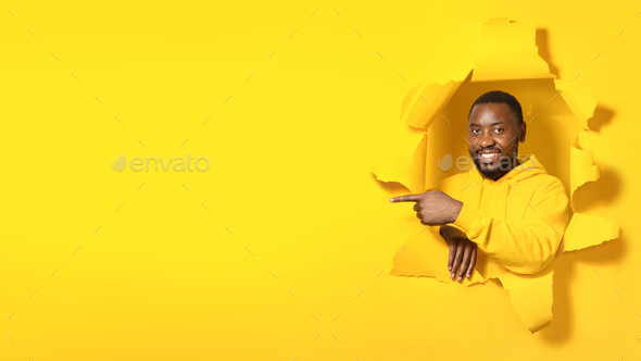 Happy black man pointing finger aside at free copy space, posing in hole in torn yellow paper