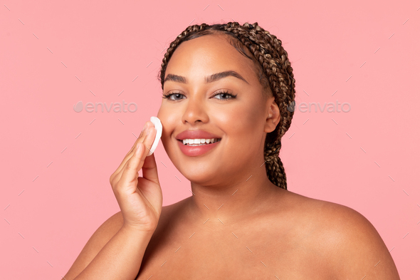 Facial skincare concept. Happy black chubby woman using cotton pad, caring for smooth skin, posing